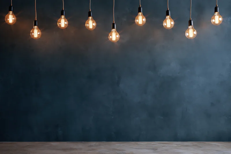 Row of glowing incandescent bulbs hanging from cords above a wooden table against a dark textured wall