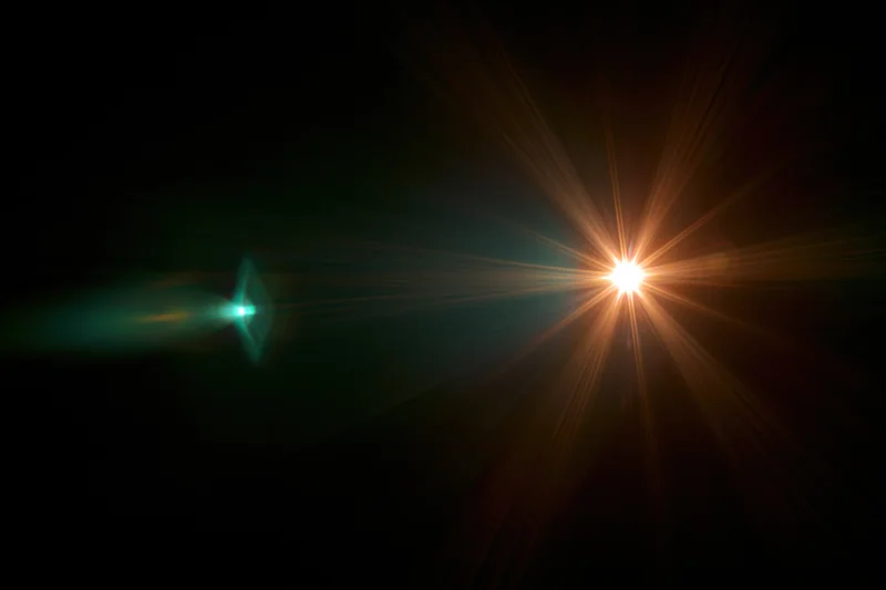 Bright point light source emitting starburst flare against dark background with green lens flare on left side