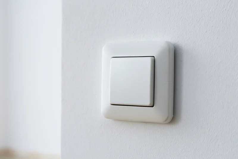 White square push-button light switch mounted on a textured white wall in an interior space