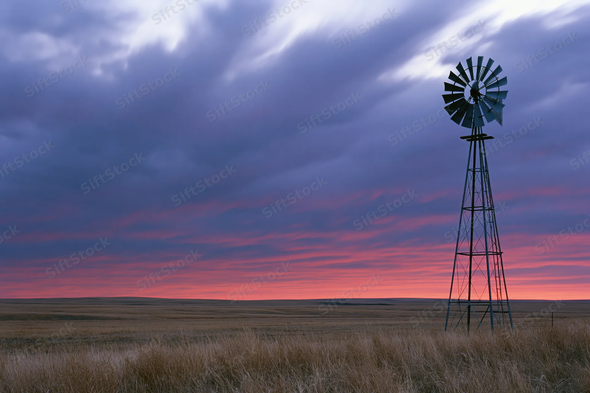 Steel farm windmill standing in open grassland at dusk with colorful sky