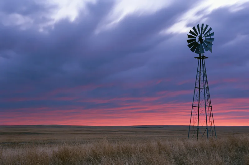 Steel farm windmill standing in open grassland at dusk with colorful sky