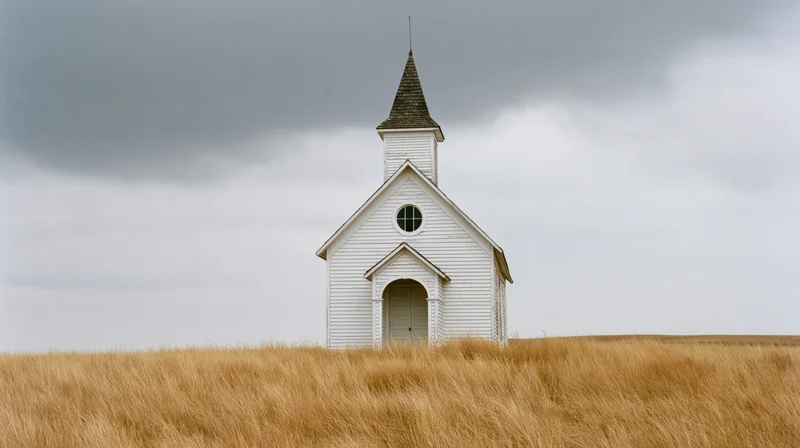 White clapboard church with pointed steeple standing alone in golden prairie grass under cloudy sky