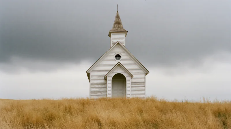 White clapboard church with pointed steeple standing alone on a golden grassy prairie under overcast sky