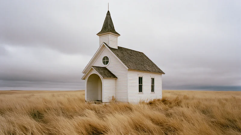 Abandoned white clapboard church with pointed steeple standing alone in a dry grassy prairie field under an overcast sky