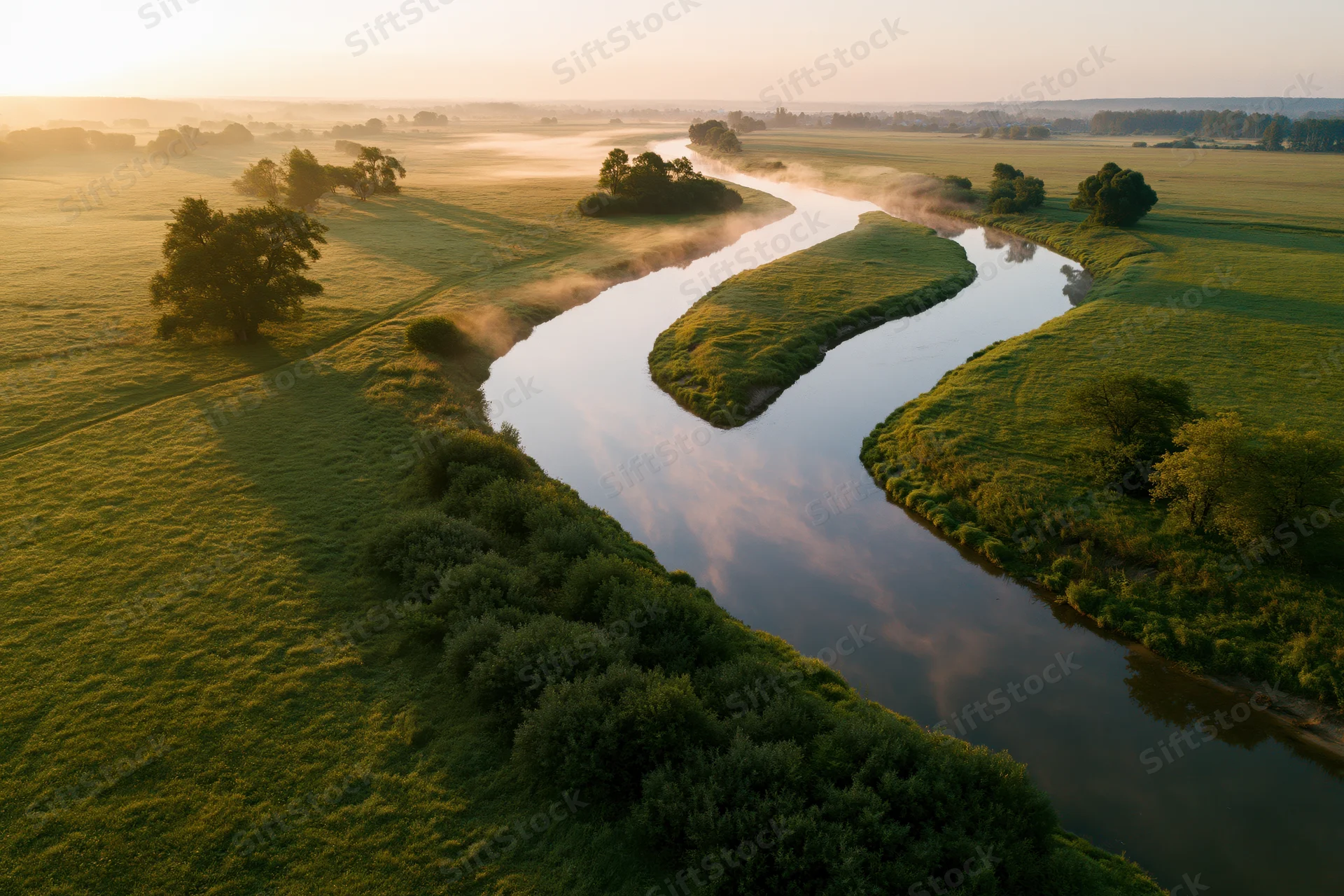 Winding river flowing through flat green meadow at sunrise with mist rising from the water and trees along the banks