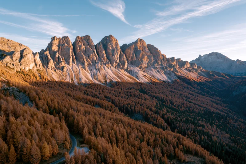 Jagged rocky peaks of the dolomites in autumn with golden larch forest and winding road below