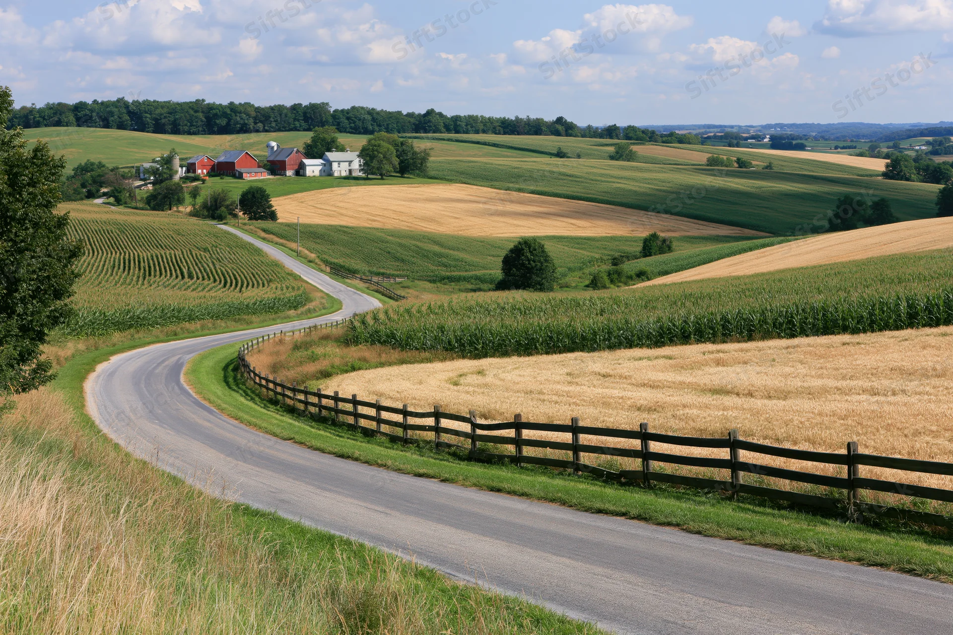 Winding asphalt road curves through rolling farmland with corn and wheat fields, barns, and wooden fences under a blue sky with clouds