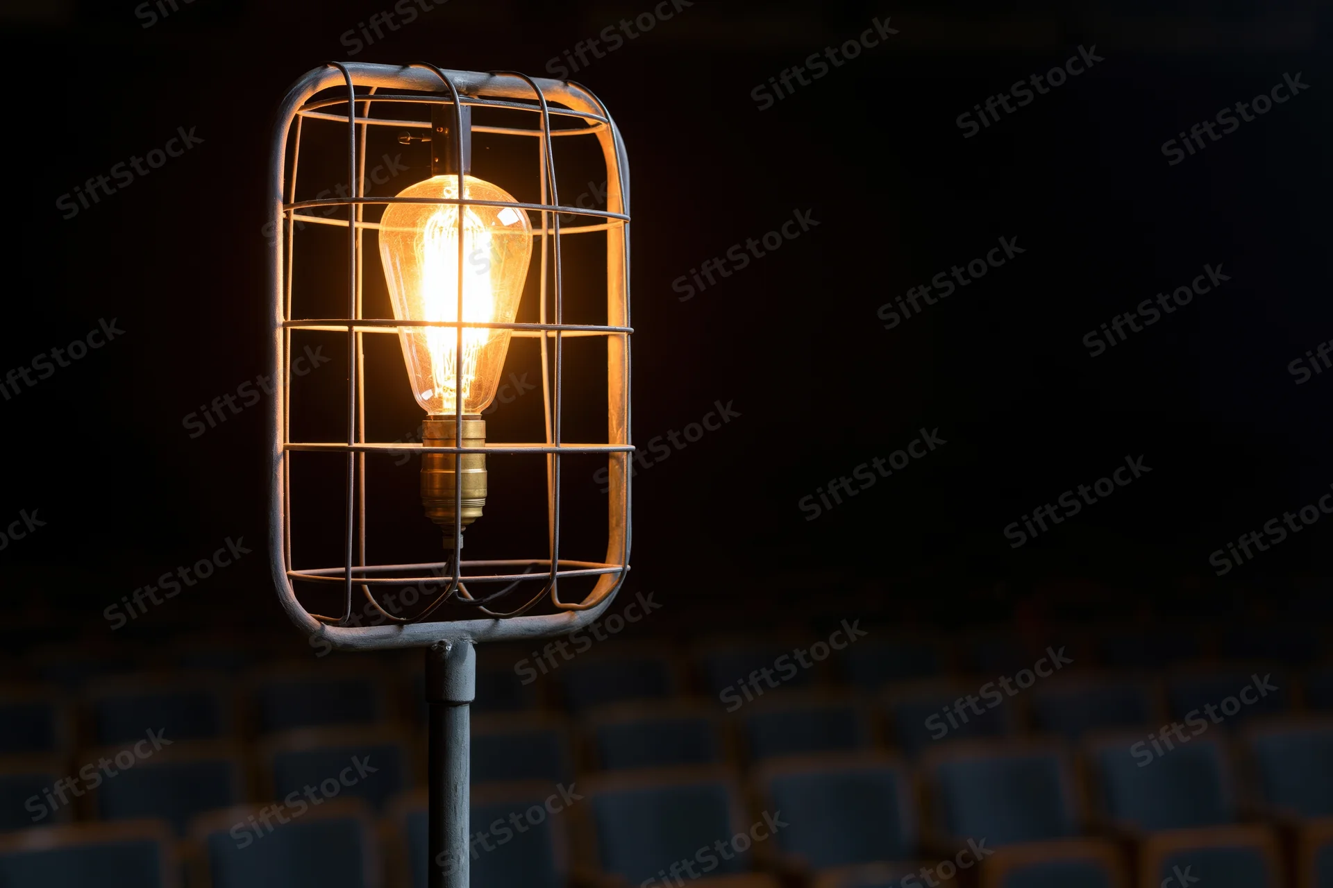 Industrial cage work light glowing warmly on metal stand in dark theater with rows of seats