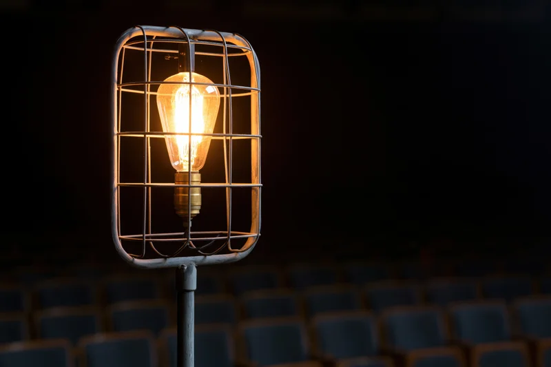 Industrial cage work light glowing warmly on metal stand in dark theater with rows of seats