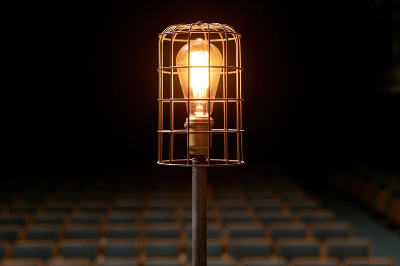 Industrial cage light glowing warmly on metal stand in dark auditorium with rows of seats