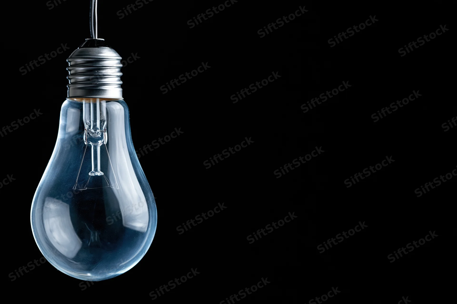 Unlit clear glass incandescent light bulb hanging from a wire against a black background with copyspace