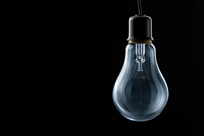Unlit clear glass incandescent light bulb hanging from a wire against black background