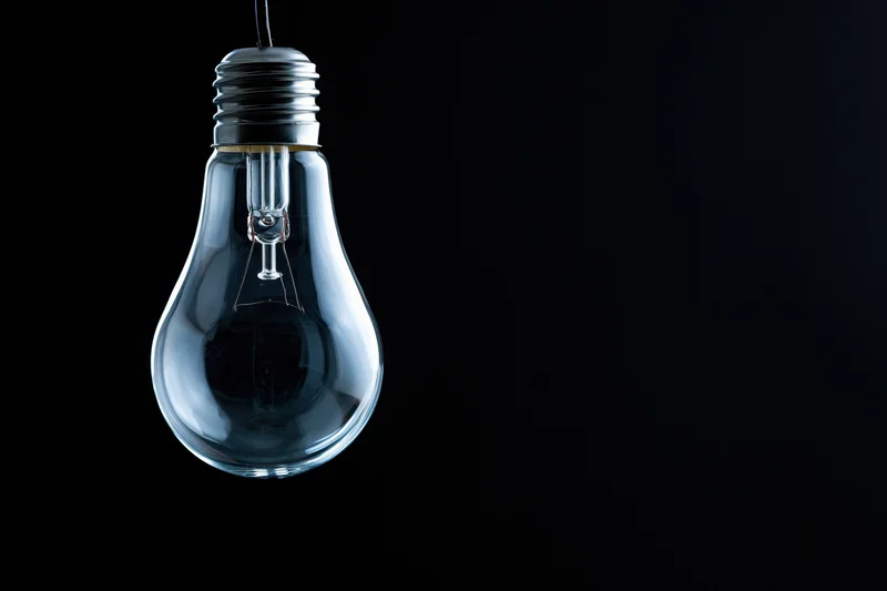 Unlit clear glass incandescent light bulb hanging from a wire against a black background