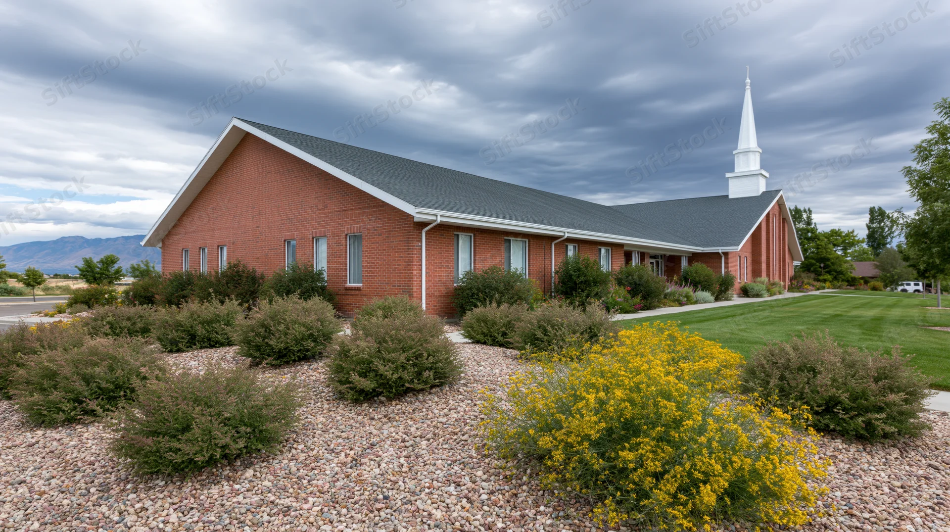 Brick lds meetinghouse with white steeple and landscaped garden under cloudy sky