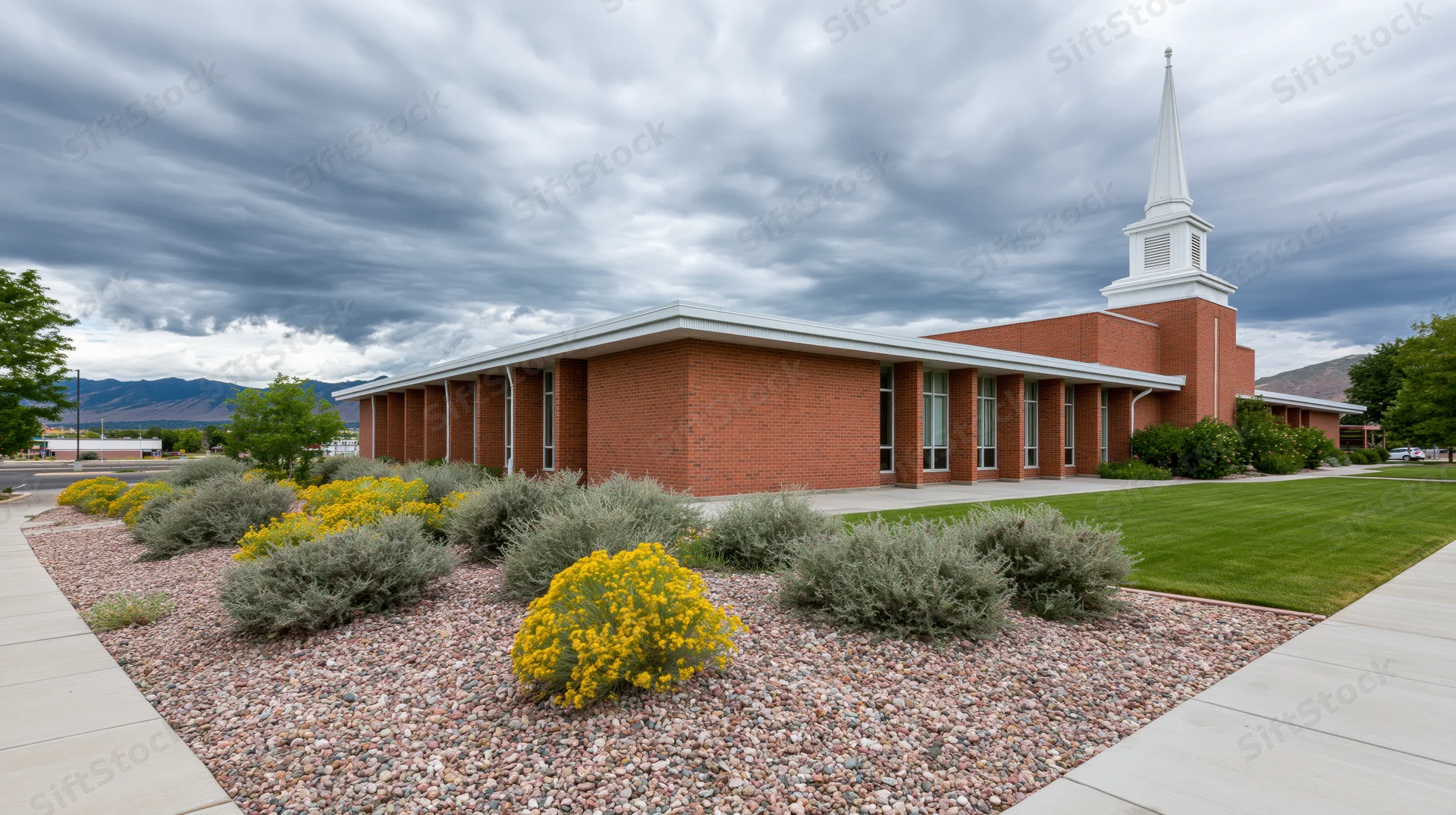 Brick lds meetinghouse with white steeple and landscaped garden under cloudy sky