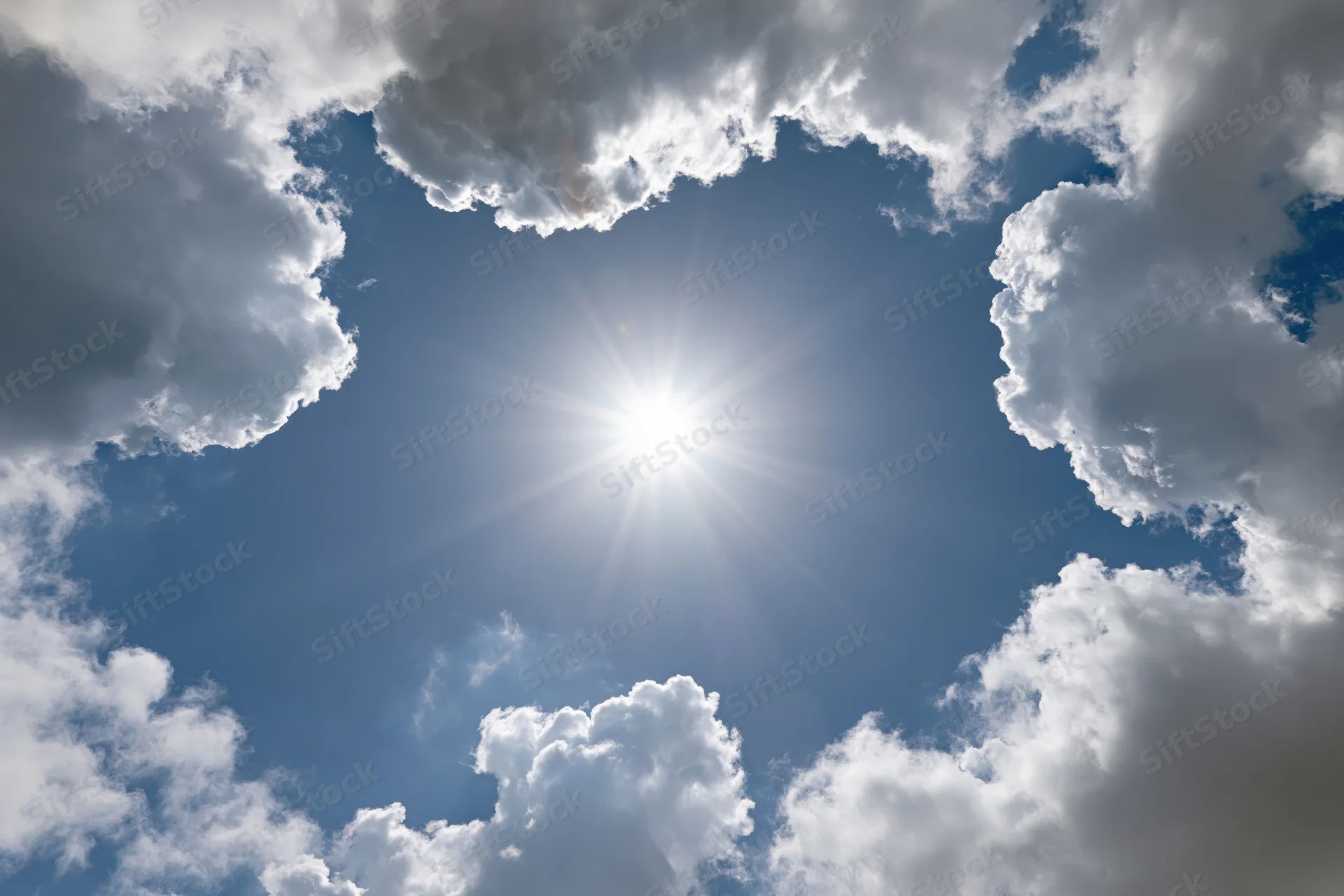 Bright sun shining through a break in white cumulus clouds against a clear blue sky