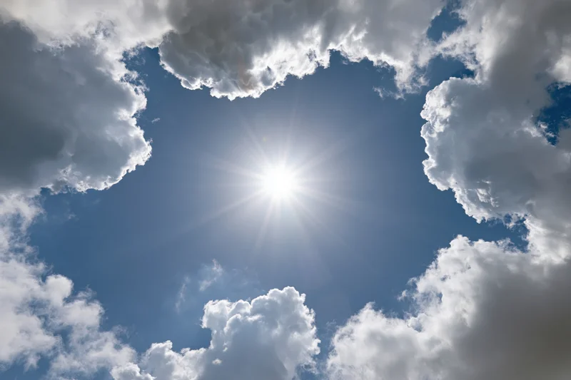Bright sun shining through a break in white cumulus clouds against a clear blue sky