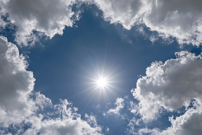 Bright sun shining through a break in white cumulus clouds against a blue sky
