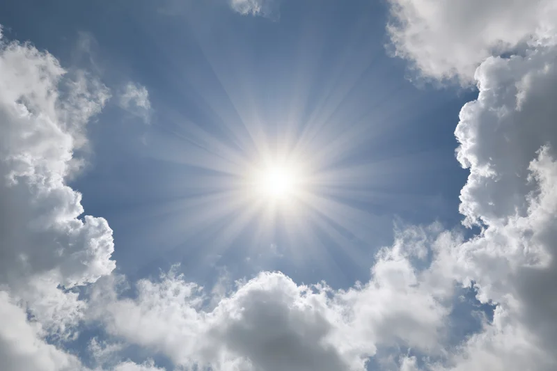 Bright sun bursting through a break in white cumulus clouds against a blue sky