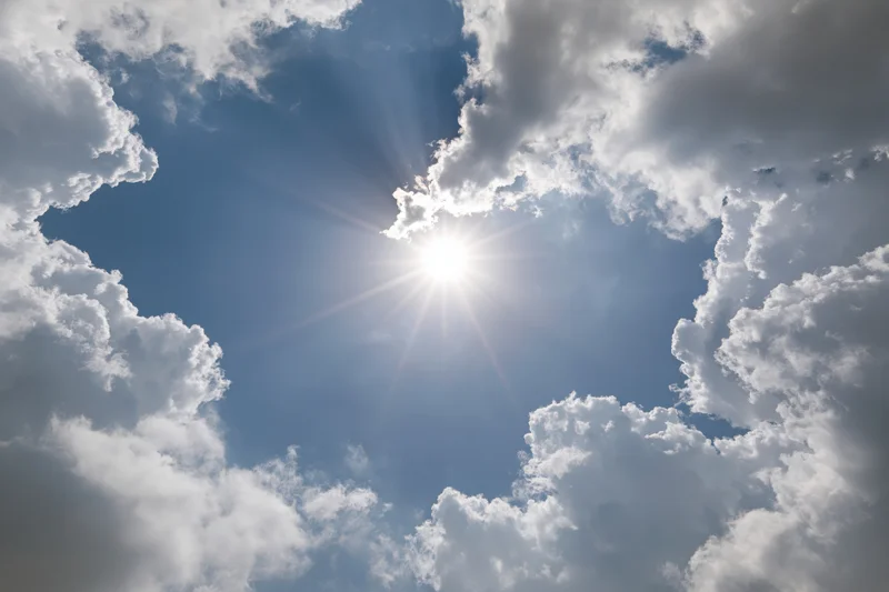 Bright sun bursting through a break in white cumulus clouds against a blue sky