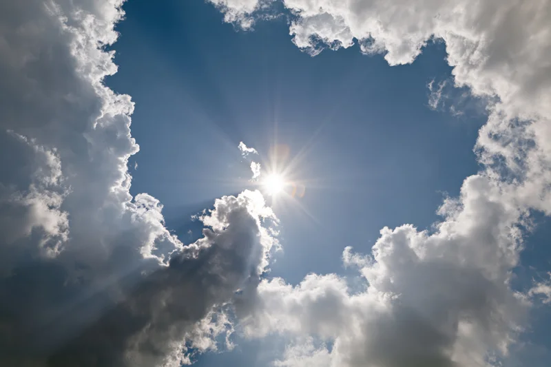 Bright sun bursting through gap in white cumulus clouds against blue sky