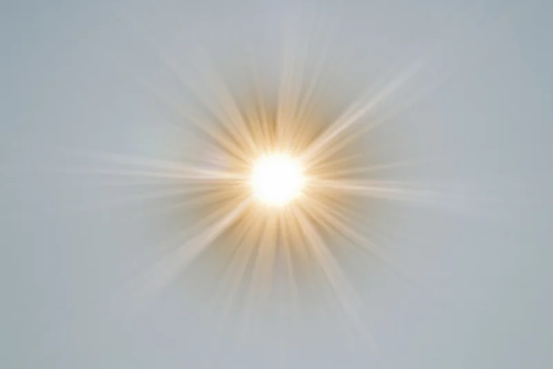 Bright sunburst lens flare radiating light rays from center against soft sky background