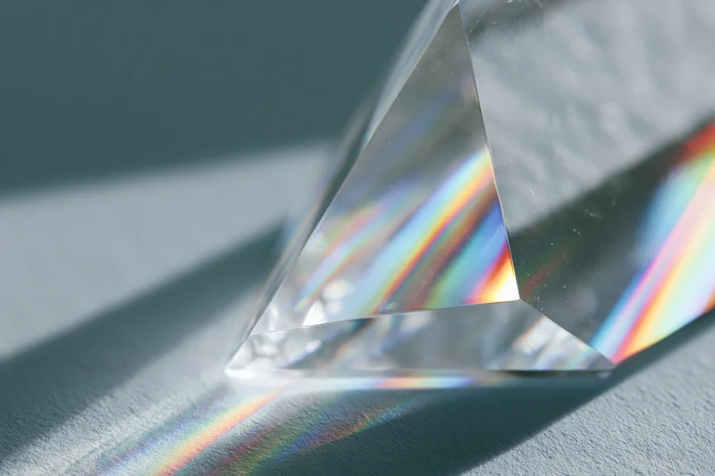 Clear glass crystal prism refracting bright light into scattered rainbow spectrum on gray surface