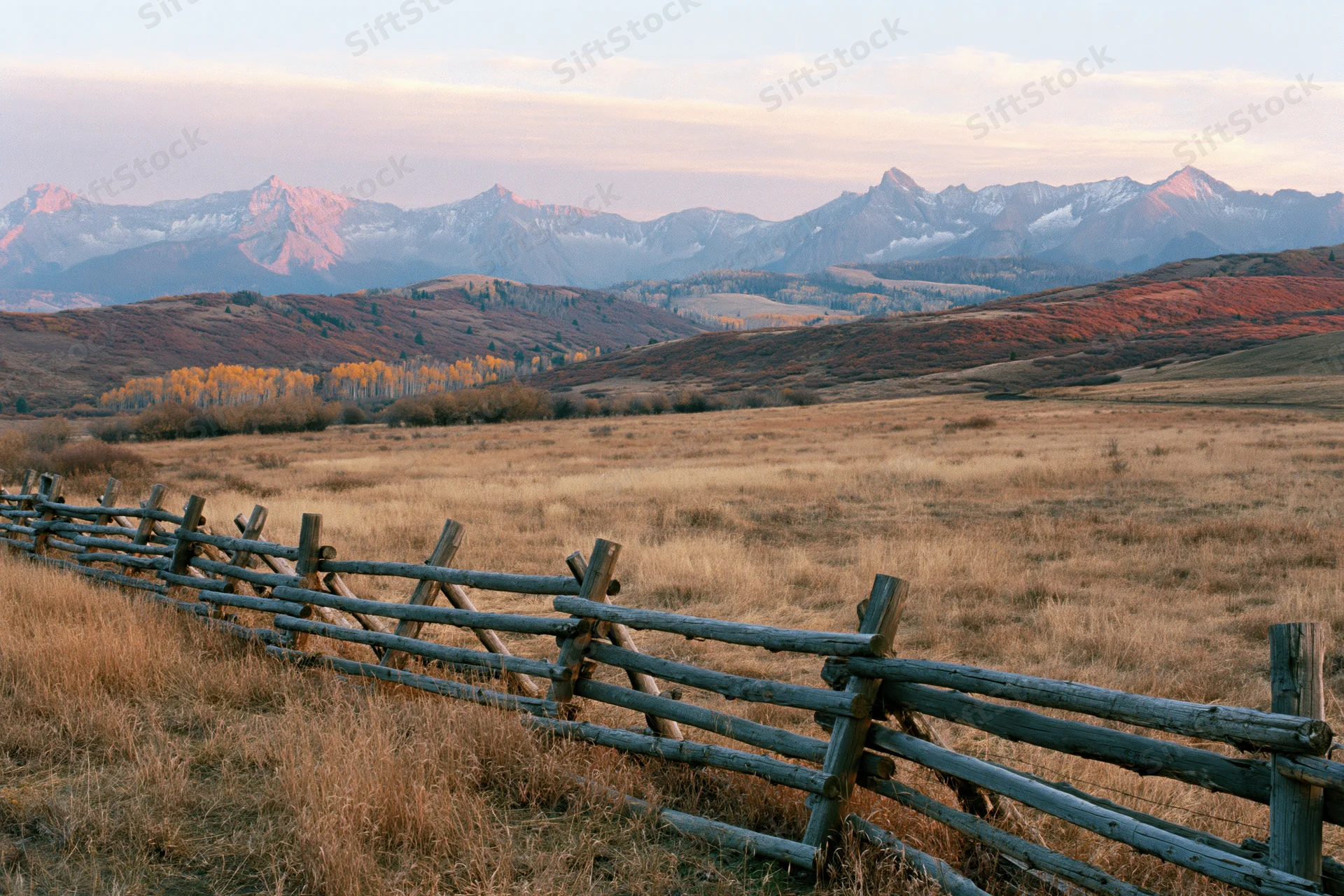 Wooden fence running through dry grassy valley with snow-capped Rocky Mountains at sunset in autumn Colorado