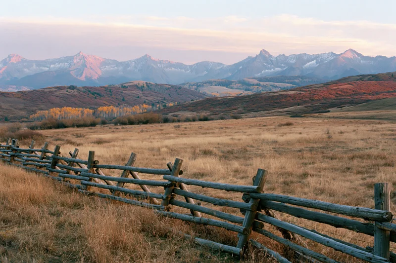 Wooden fence running through dry grassy valley with snow-capped Rocky Mountains at sunset in autumn Colorado
