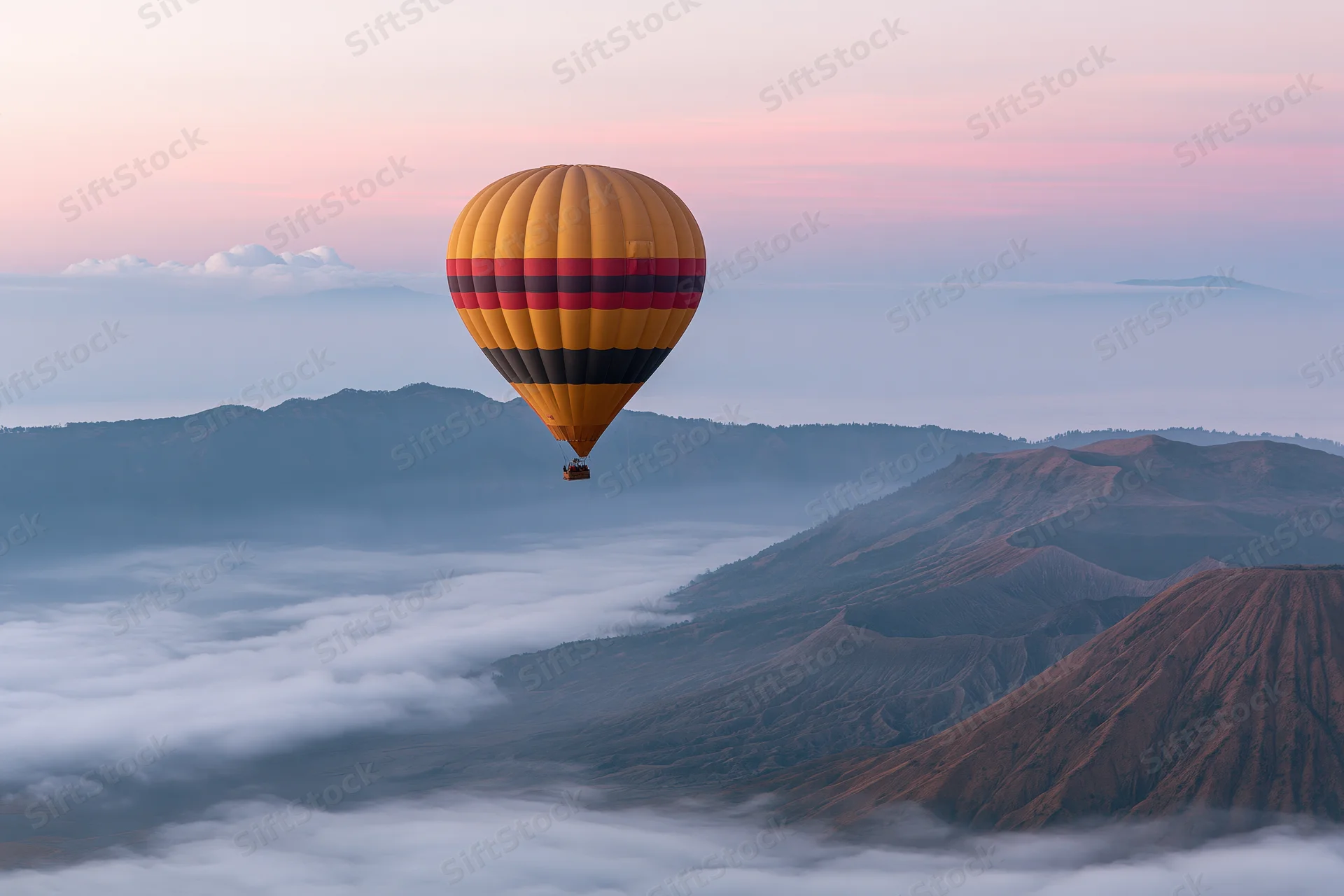 Colorful striped hot air balloon drifting above volcanic mountains in misty valley at sunrise