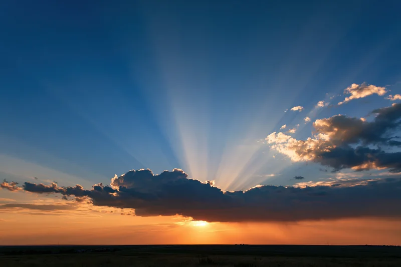 Crepuscular rays bursting upward from clouds at sunset over open field