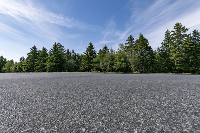 Empty asphalt road surface stretching toward a treeline of evergreen trees under a blue sky with wispy clouds