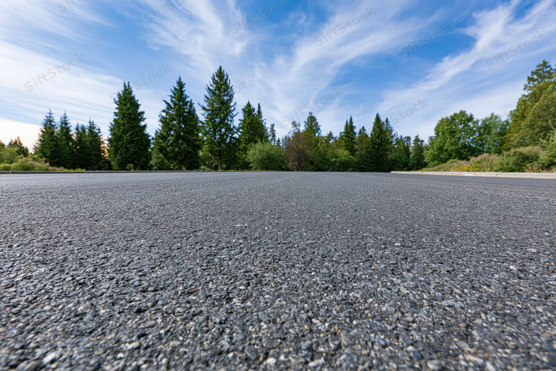 Empty asphalt road surface stretching toward a treeline of evergreen trees under a blue sky with wispy clouds
