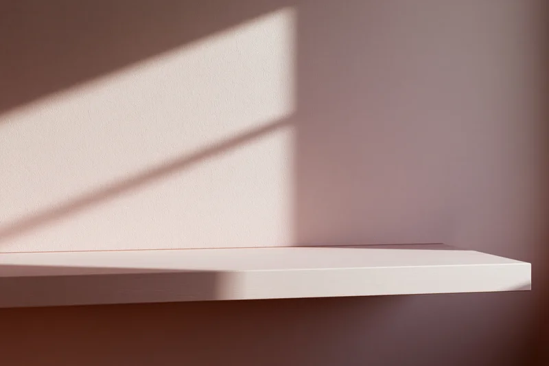Empty white shelf mounted on a flat matte pink wall with sunlight casting diagonal shadows across its surface