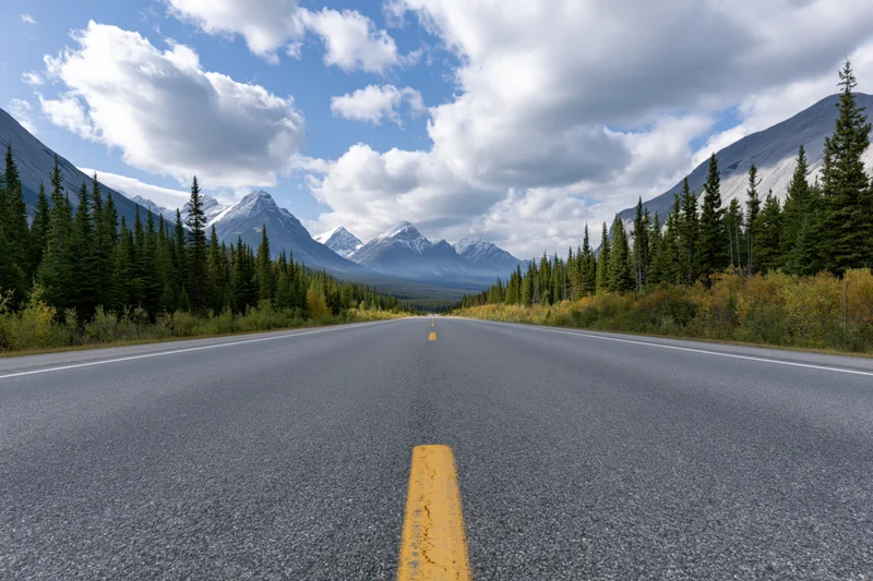 Empty two-lane asphalt highway with yellow center dashes stretching toward snow-capped mountains flanked by pine forest and autumn foliage