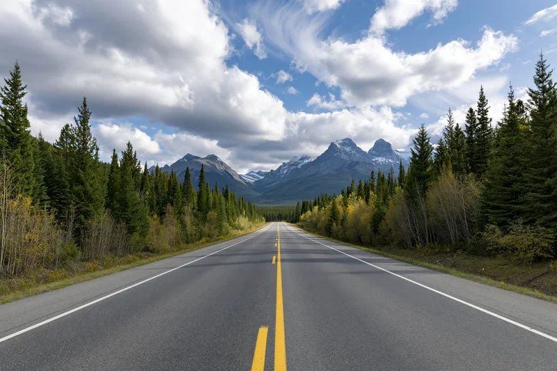 Empty two-lane asphalt highway with yellow center dashes stretching toward snow-capped mountains framed by evergreen forest and autumn foliage under a partly cloudy sky