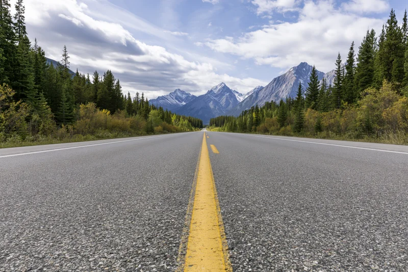 Empty two-lane asphalt highway with yellow dashed centerline leading toward snow-capped mountains framed by evergreen forest