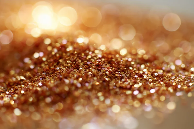 Scattered gold and bronze glitter particles on a textured surface with bokeh lights in the background