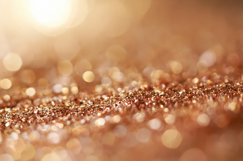 Scattered gold and bronze glitter particles on a textured surface with bokeh light reflections in the background