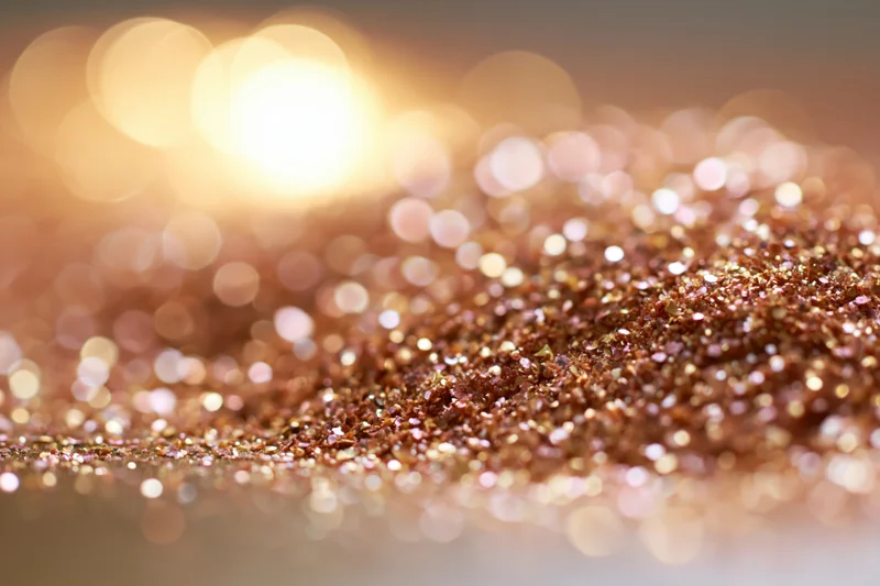 Scattered gold and bronze glitter particles on a reflective surface with bokeh lights in the background