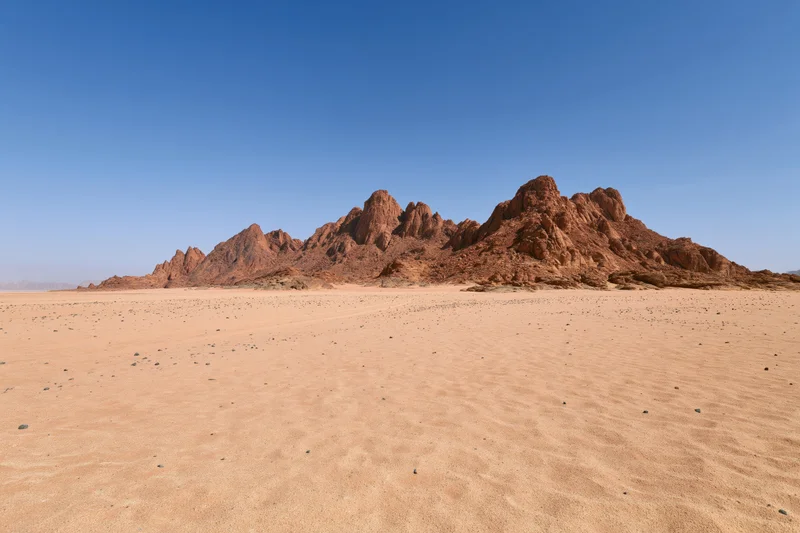 Flat sandy desert floor stretching toward rugged sandstone rock formations under clear blue sky