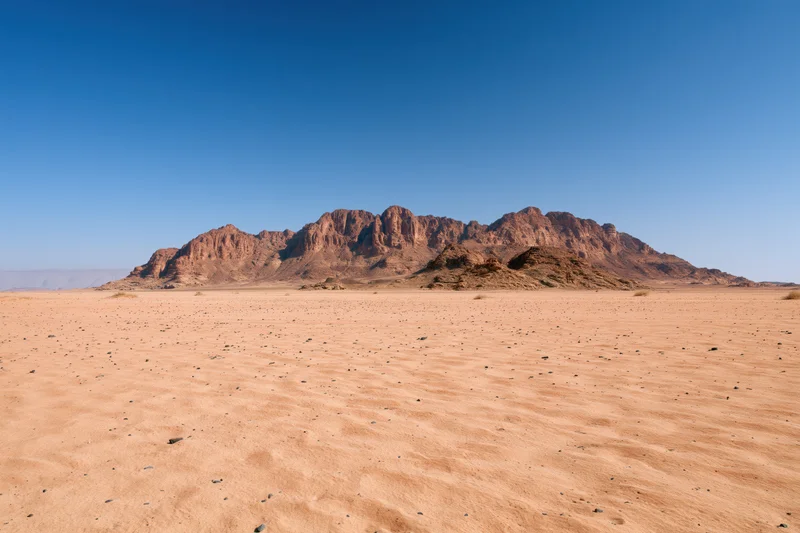 Flat desert floor stretching toward rugged sandstone mountains under clear blue sky