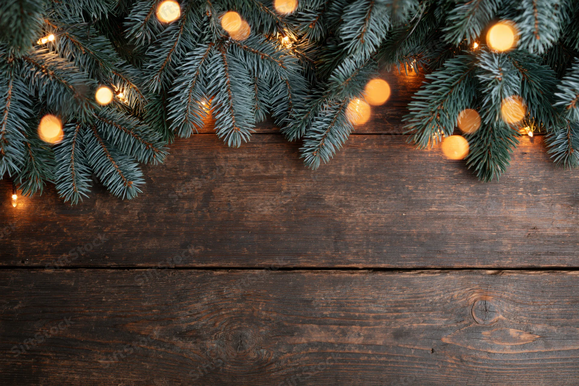 Fir and spruce branches with warm string lights arranged along the top edge of a dark wooden surface, leaving copyspace below