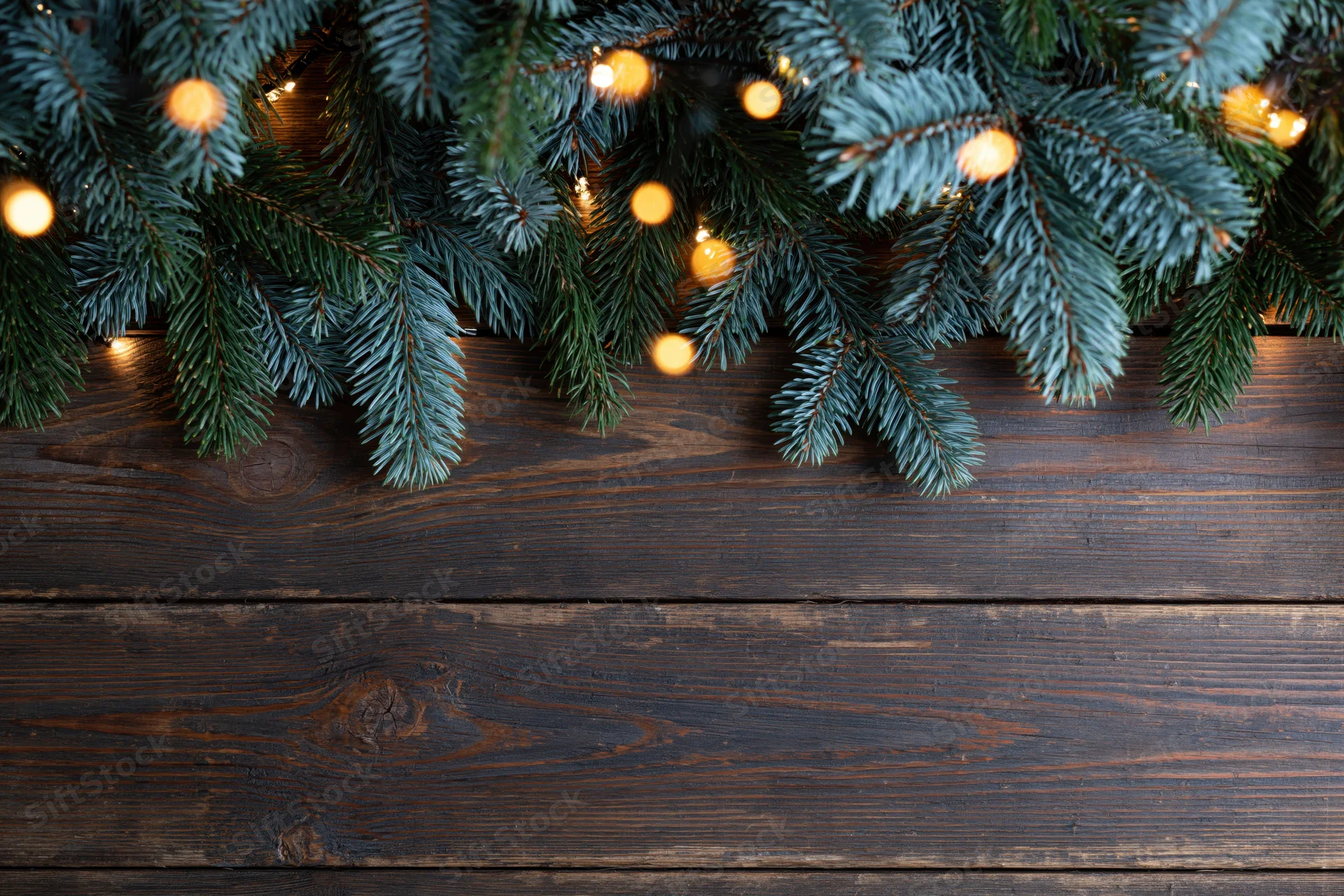 Fir and spruce branches with warm string lights arranged along the top edge of a dark wooden surface