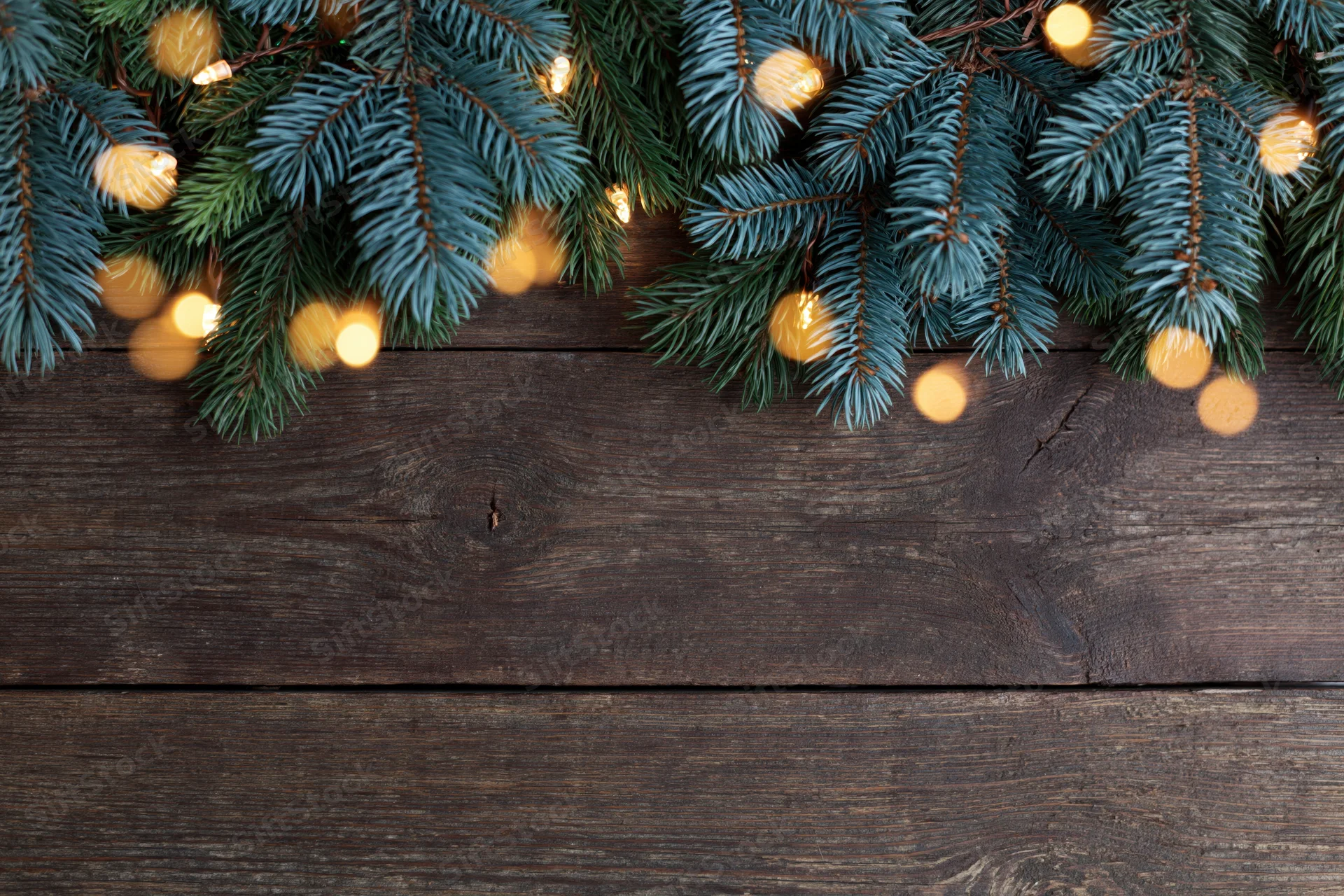 Fir and spruce branches with warm lights arranged along the top edge of a dark wooden surface, leaving copyspace below