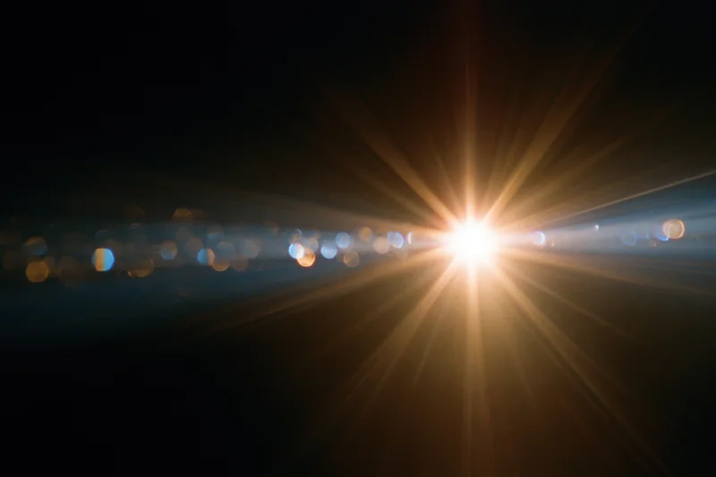 Intense lens flare from a bright point light source radiating rays across a dark background with bokeh lights