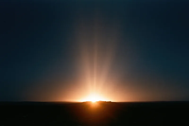Intense sun rising over horizon casting long rays across dark landscape at dawn