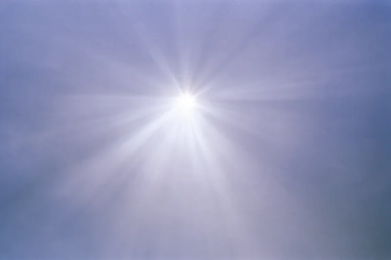 Intense sun flare bursting from upper center of frame against blue sky
