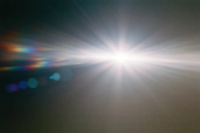 Intense white light source blazing against a near-black background with lens flare and rainbow spectrum streaks