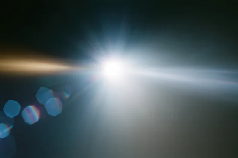Intense white light source blazing against a near-black background with lens flare and rainbow bokeh patterns